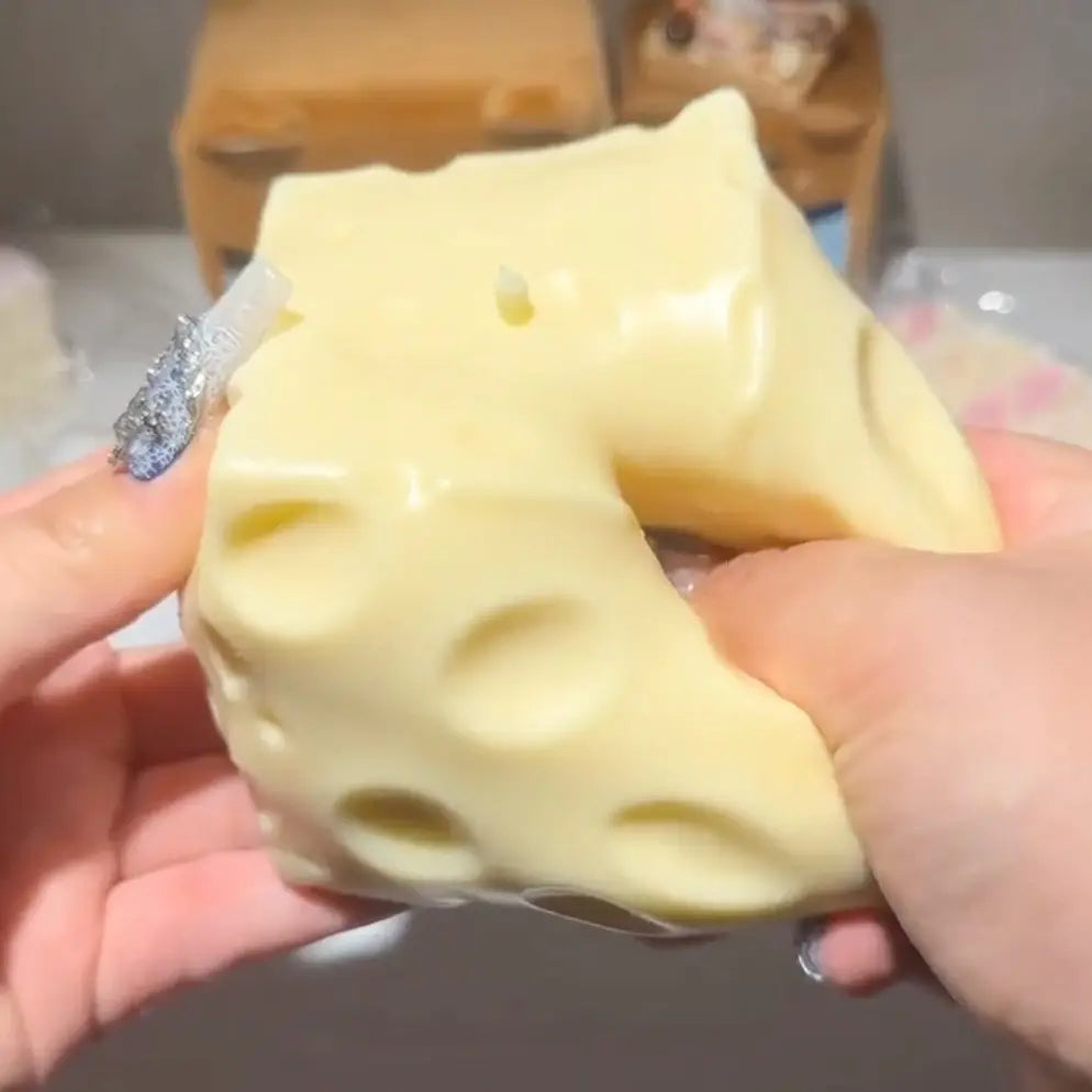 A pale yellow, cheese-shaped squishy toy being held and squeezed by two hands. The toy features a molded surface with several circular holes resembling Swiss cheese.