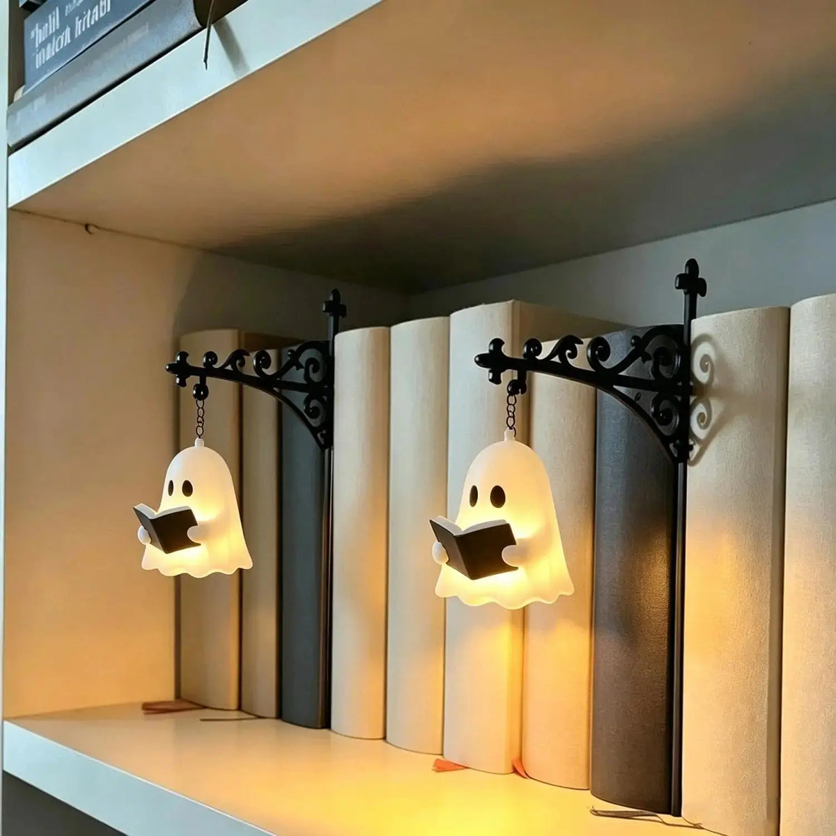 Two decorative wall sconces shaped like glowing ghost books hanging from black wrought-iron brackets, mounted on a shelf in front of a row of tall books.