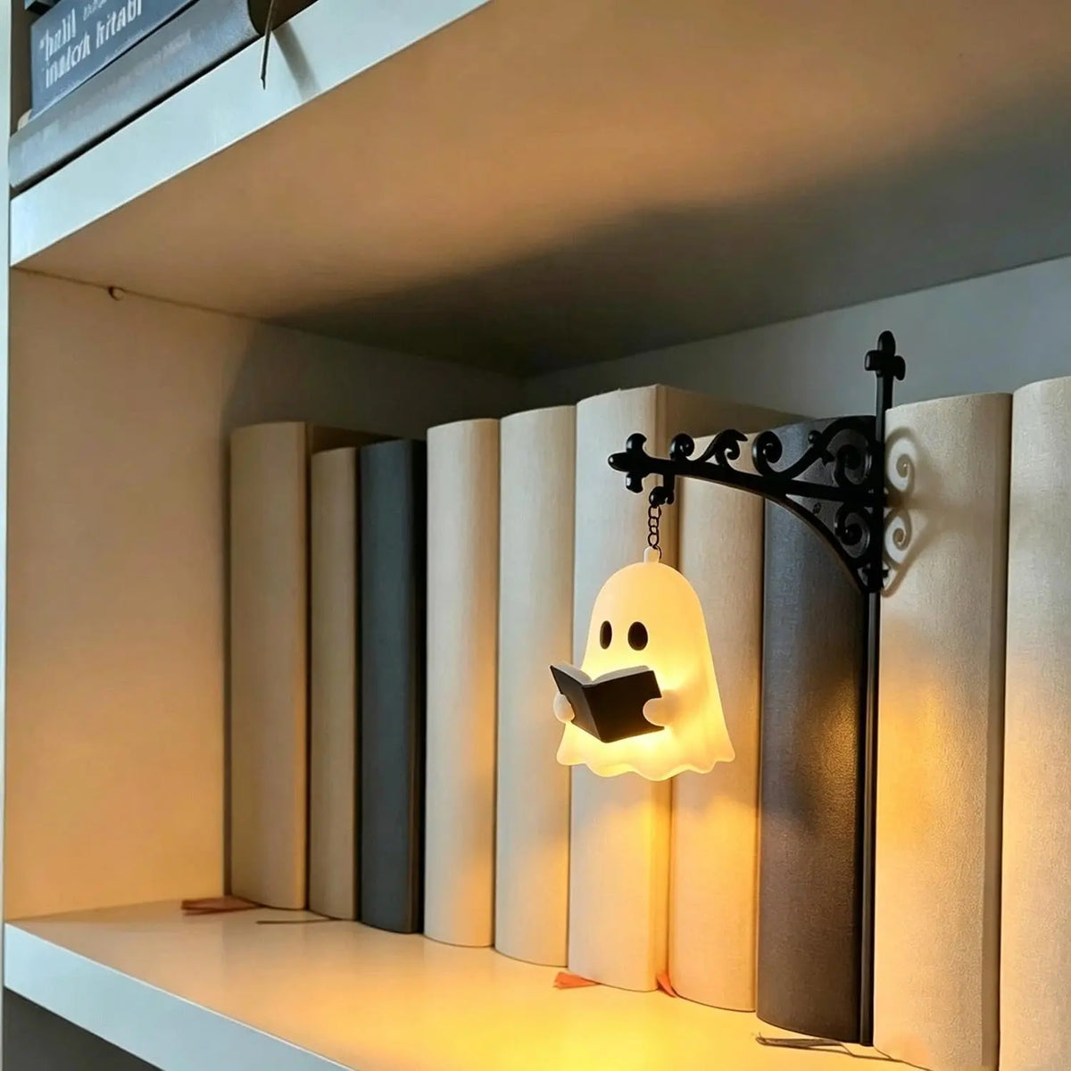 A ghost-shaped wall lamp glowing warm white, holding an open book, mounted on a black wrought-iron bracket on a shelf between books.